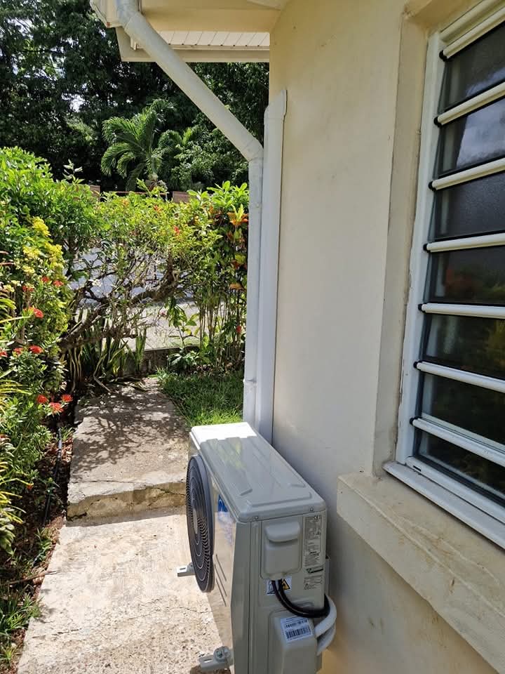 Installation Midea 9k BTU – Ducos – climatisation Martinique Installation Midea 9 000 BTU à Ducos : unité extérieure posée au sol sur une allée en béton, adossée à la façade et abritée par l'avant-toit – Ducos, Martinique – LD CLIM 972